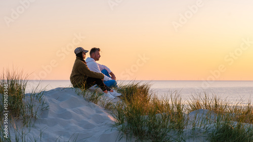 Εκτύπωση καμβά A couple sits on a sandy dune, surrounded by tall grass, admiring the vibrant sunset over the calm waters of the beach in Denmark