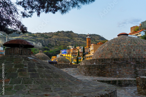 Stone domes of sulfur baths, houses with carved balconies, Tbilisi Juma Mosque and part of the wall of Narikala fortress. Hill and blue sky in the background photo