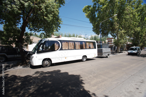 A small white passenger bus is parked in the shade of large trees in a rural area, ready for a trip. The beginning of the tourist season