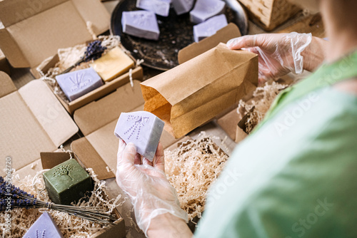 Person wearing gloves places lavender soap into a kraft bag among handmade skincare products. Handmade soap process, lavender skincare, indie soap maker, ethical manufacturing