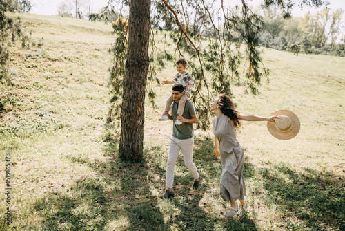 Playful family moment in a sunny park, with mother holding straw hat and father carrying son on shoulders.