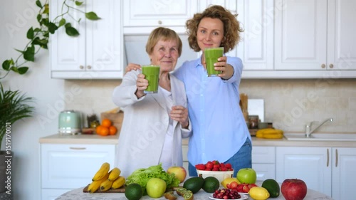 Elderly woman and daughter smiling while holding green smoothies in kitchen filled with fresh fruits. Emphasizing healthy lifestyle and family bonding