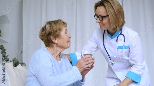 Compassionate nurse providing attentive hydration support, gently helping senior woman drinking water during medical care interaction in home healthcare setting