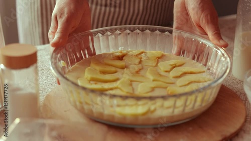 Hands hold a glass baking dish filled with apple slices arranged in batter, ready for baking a homemade dessert in a sunlit kitchen.