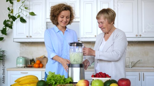 Happy family preparing healthy green smoothie with blender in kitchen, senior mother and adult daughter enjoying time together while making fresh organic vegetable juice