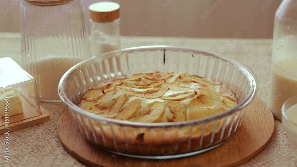 A freshly baked apple cake in a glass dish sits on a wooden board, surrounded by butter, milk, and sugar, on a rustic, sunlit table.