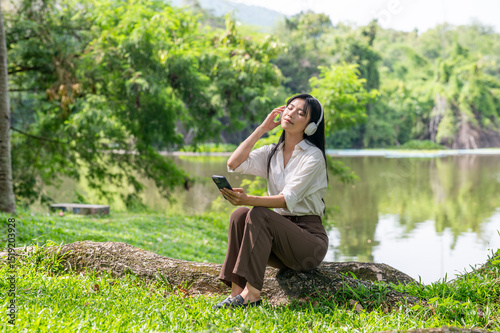 Asian woman in white shirt with headphone closing eyes listening to music sitting on a big tree root