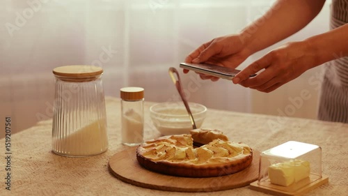 A person uses a smartphone to photograph a freshly baked apple cake on a wooden board, surrounded by baking ingredients in a rustic kitchen setting.