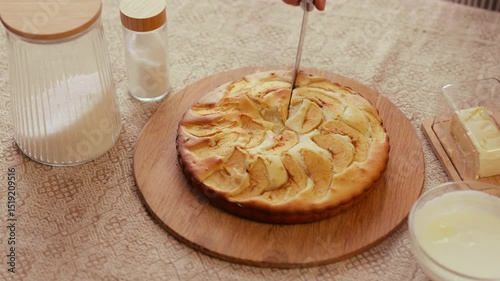 A person uses a pie server to lift a slice of freshly baked apple cake from a round wooden board, surrounded by baking ingredients.