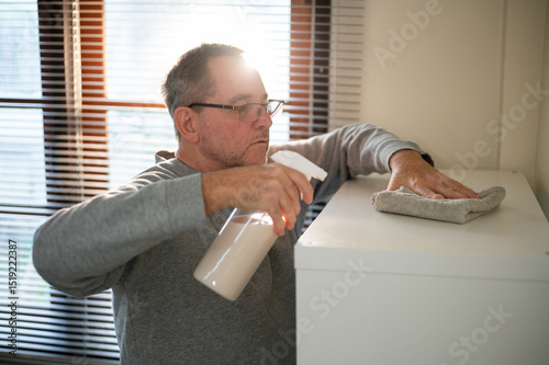 Old white caucasian man holding cleaning foggy spray and cloth rag wiping dust off shelf in his room