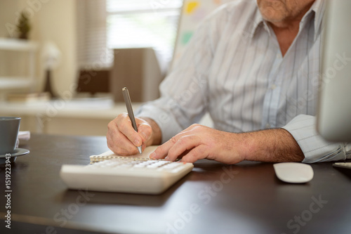 Close up of an old man hands holding pen writing in notebook with calculator aside on working table.