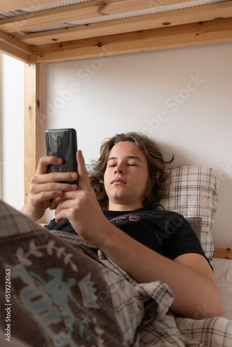 A young man checks his smartphone in bed after waking up in the morning. He is scrolling through social media or news, starting his day connected to the internet and online content.
