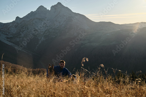 Photography Man resting in tall grass with two dogs - german and australian shepherds - at sunset, with Komovi mountains in background