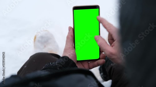 Young man sitting and holding smartphone with green screen outdoors in winter. Vertical mockup of mobile phone. Guy using phone with blank green display background. Smartphone with chromakey. Close-up