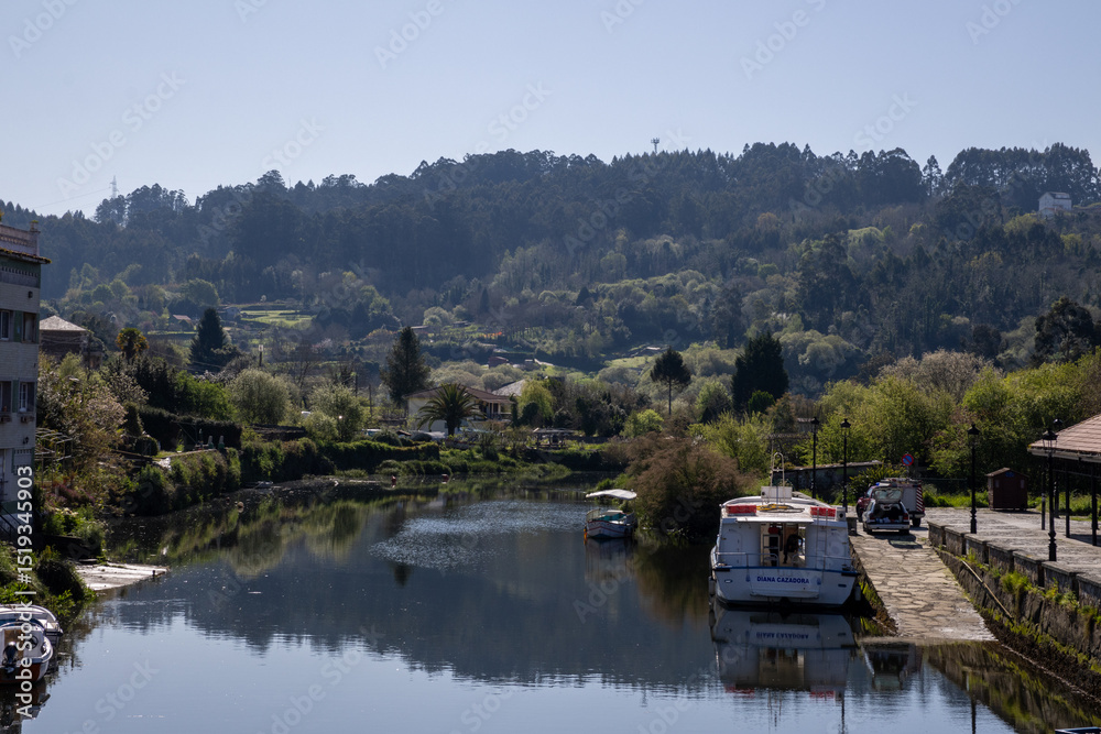Fototapeta premium Scenic view of Ponte Vella bridge over Mandeo River in Betanzos, Galicia, Spain, surrounded by lush greenery and historic architecture.
