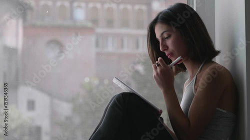 A young woman sits quietly on a window ledge, writing a heartfelt goodbye letter to her partner. The moment is tender and filled with emotion.