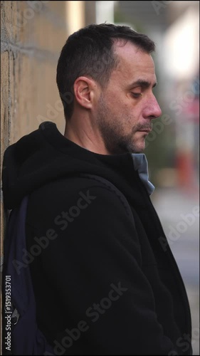 Vertical: A young man stands leaning against a wall outside, looking down in silence with visible sadness
