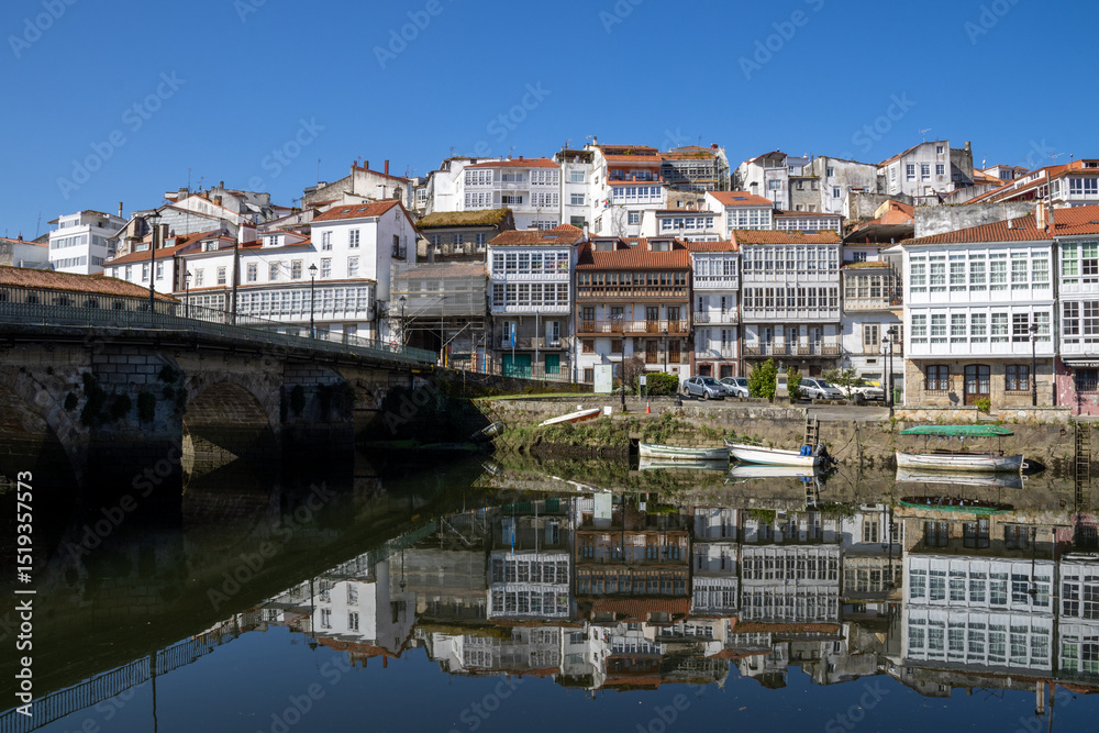 Obraz premium Scenic view from Praza a Enrique Ivntiga Eira Vella, Betanzos, Galicia, with riverside white homes and boats reflecting on the peaceful Mandeo River.