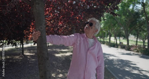 Energetic senior from Madrid in pink shirt playfully interacts with a tree, enjoying nature with youthful enthusiasm during a sunny walk in a peaceful urban park.