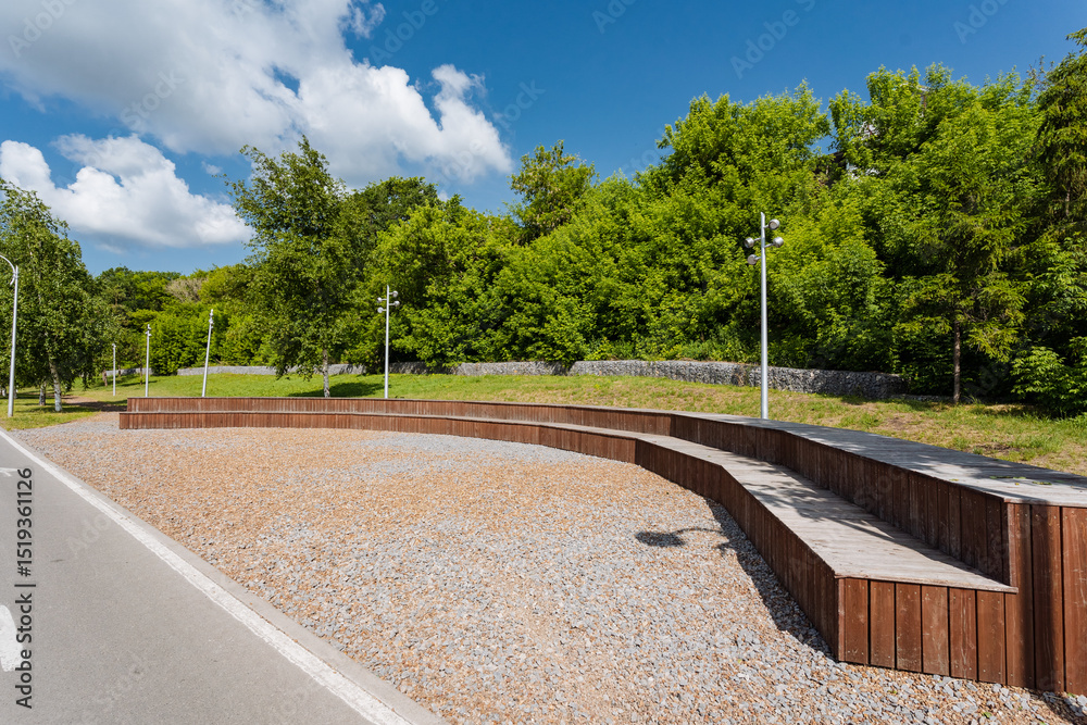 Obraz premium In a pleasant park, a sturdy wooden bench can be found situated right alongside the side of a road, providing a resting spot for visitors enjoying their surroundings and natures beauty