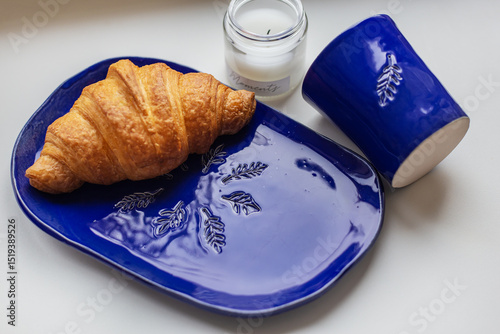 Handmade blue clay plate with croissant on it. Vase with wheat and candles. photo