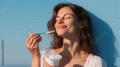 Woman enjoys eating Yogurt outdoors on a bright, sunny day against a blue wall, feeling pleased with her healthy diet