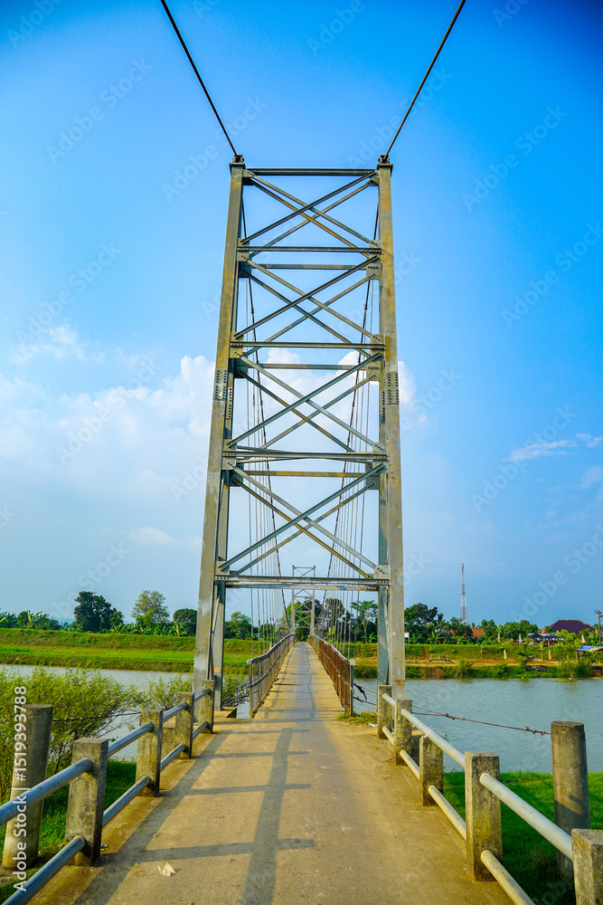 Fototapeta premium a towering suspension bridge tower with an iron frame structure against a clear sky backdrop for advertising space. A steel suspension bridge spans a river flanked by greenery under a bright blue sky.