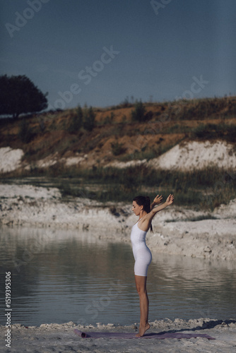 Woman in white sportswear sitting on yoga mat by lake holding foot in stretching pose. Outdoor fitness and flexibility training concept. Design for poster, banner, greeting card, invitation. Side view