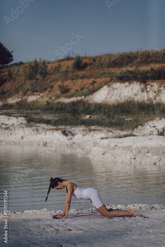 Woman in white sportswear kneeling on yoga mat by lake in stretching position. Outdoor fitness and healthy lifestyle concept. Design for poster, banner, greeting card, invitation. Side view