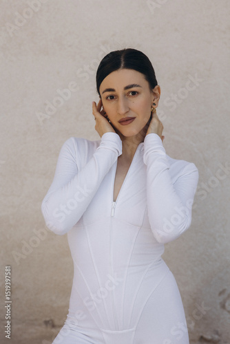 Woman in white zip-up bodysuit touching her face. Studio fashion portrait on beige textured background. Beauty and style concept. Design for poster, banner, greeting card, invitation. Front view