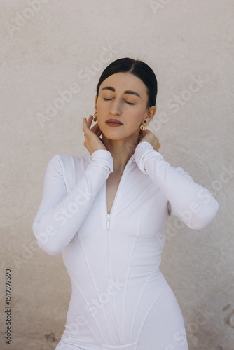Woman in white zip-up bodysuit with closed eyes touching neck. Studio fashion portrait on beige textured background. Beauty and style concept. Design for poster, banner, greeting card, invitation. 