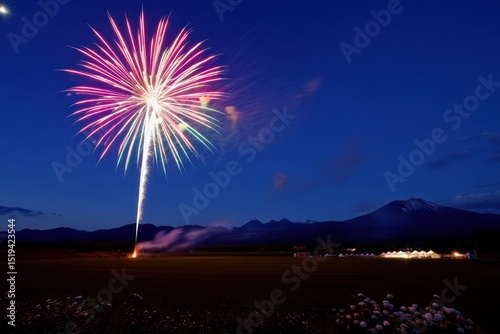 Colorful fireworks exploding in the night sky over a mountainous landscape with bright tents glowing in the distance and flowers in the foreground