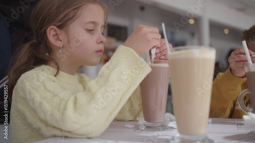 Girl in yellow sweater adding sugar from small sachet into tall glass of chocolate drink at cafe table. Focused expression on face. Cozy indoor setting with soft light and relaxed atmosphere