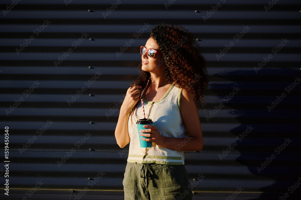 Obraz premium woman with coffee cup enjoys sunshine outdoors wearing sunglasses and casual outfit. sunlight and shadows create dramatic contrast on textured background inspiring summer vibes