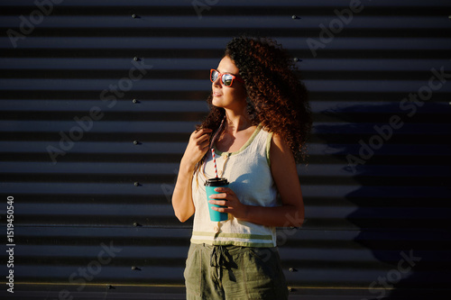 woman with coffee cup enjoys sunshine outdoors wearing sunglasses and casual outfit. sunlight and shadows create dramatic contrast on textured background inspiring summer vibes photo