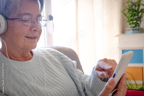 Relaxed senior woman sitting in home living room using mobile phone. Smiling grandmother wearing headphones listening to music