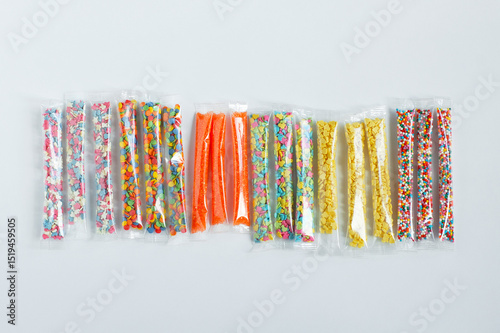Multi-colored confectionery sprinkles in individual transparent packaging in a row on a white background.