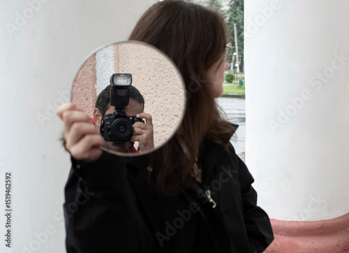 A young woman holding a mirror in which a photographer with a camera is reflected