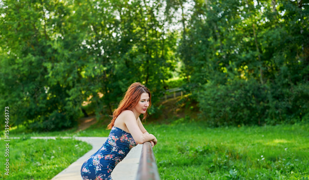 Naklejka premium Woman in blue dress leaning on bridge railing in park. Relaxation and rest concept as woman enjoying her time outdoors. Lush green surroundings. Girl has beautiful body.