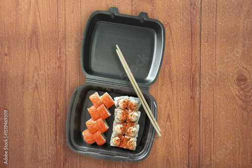 Sushi rolls in gray packaging and chopsticks on brown wooden background.