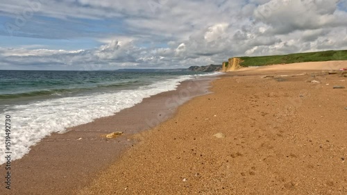 Slow Motion footage of the waves crashing up onto a beach on the Jurassic Coast, Dorset. The footage is low and moves backwards with amazing cliffs in the background and a sky full of clouds above.