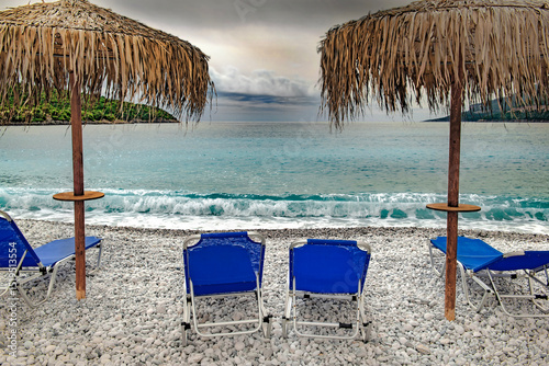 Fototapeta Naklejka Na Ścianę i Meble -  straw umbrella on a beach of white pebbles  with lounge chairs in front of a beautiful sea under cloudy sky in Peloponnese, Greece