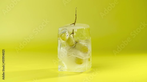 Grape in an ice cube, close-up. Time lapse of the ice melts and a bunch of green grape appears. Frozen fruit. Yellow background.