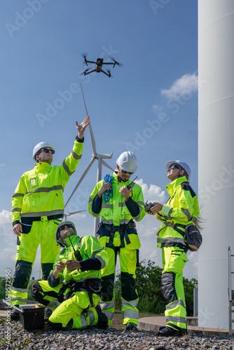 Maintenance engineer team standing at windmills at wind turbine farm. Group of people wear safety helmet and uniform working at alternative renewable energy wind station. Sustainable energy technology