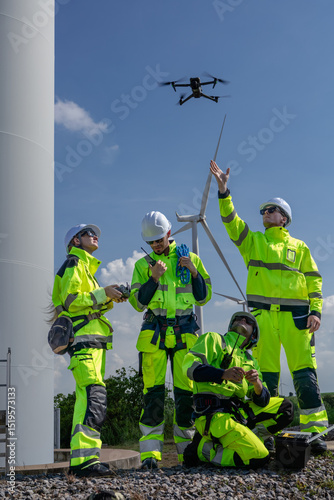 Maintenance engineer team standing at windmills at wind turbine farm. Group of people wear safety helmet and uniform working at alternative renewable energy wind station. Sustainable energy technology