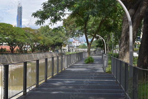 Scenic Riverside Walkway in Brickfields, Kuala Lumpur, Malaysia. The pathway runs parallel to the river, with tall buildings and tropical trees in the background.