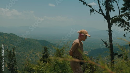 Young beautiful girl in cap walks on hill and admires majestic view of green mountains and valleys under blue summer sky. Slow motion.