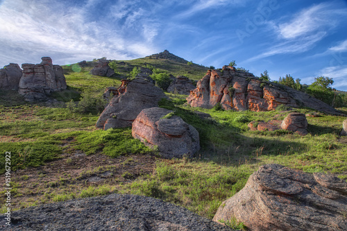 Rocks in the mountains against the background of green vegetation of grass and forest and trees under light feathery clouds. Beautiful summer landscape at sunset