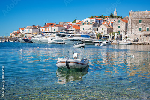 A picturesque coastal town of Primosten in Croatia features clear turquoise water, moored yachts, stone houses with red rooftops, and a peaceful Mediterranean atmosphere under a bright blue sky. photo