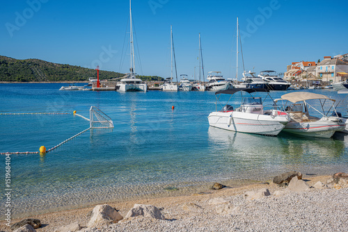 Fototapeta Naklejka Na Ścianę i Meble -  A tranquil coastal scene featuring clear turquoise waters, anchored yachts, and a green hillside under a bright blue sky in Primosten, Croatia.